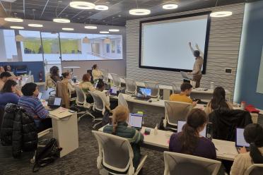 Attendees to our software carpentry workshop in the Darwin training suite at EI. Researcher Nicola Soranzo stands at the front of the room pointing at the large display screen. Participants sit at rows of curved desks around the screen.